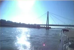 Aft-looking port side camera view showing the river, cable-stayed bridge pylon, and port railing.