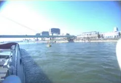 Aft-looking starboard side camera view showing the bridge, right riverbank buildings, and starboard deck.