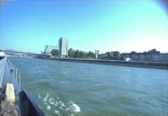 Forward-looking port side camera view showing the port gunwale, river surface, and shoreline buildings.
