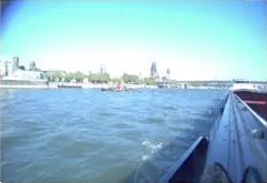 Forward-looking starboard side camera view along the hull toward the riverbank and distant bridge.